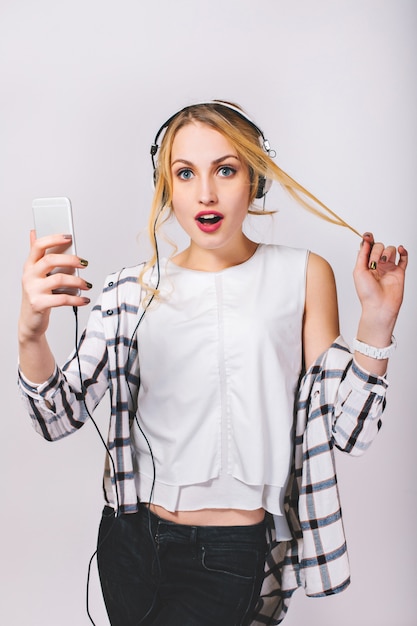 Aantrekkelijke jonge blonde vrouw met grote witte koptelefoon luisteren naar muziek op smartphone. Leuk meisje wat betreft haar haar. Verrast grote blauwe ogen, geopende mond. Geïsoleerd.