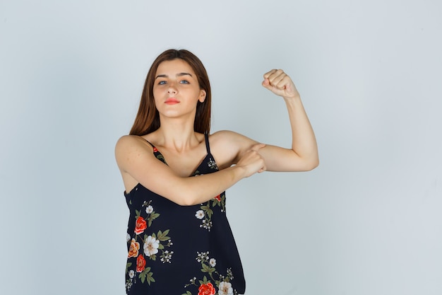 Aantrekkelijke jonge dame die de spieren van de arm in blouse toont en er trots uitziet. vooraanzicht.