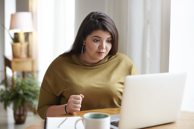 Aantrekkelijke jonge donkerharige vrouw met bochtige lichaam met behulp van generieke laptop voor extern werk, koffie drinken, scherm kijken met geconcentreerde gerichte gezichtsuitdrukking. Technologie en levensstijl