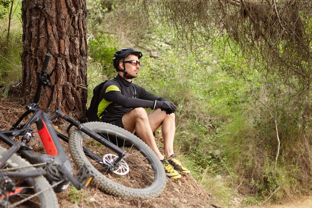 Aantrekkelijke jonge Europese rijder in beschermende kleding zittend op de grond op boom, overweegt verbazingwekkende wilde natuur om hem heen terwijl hij rust na intensieve fietstraining in het bos op zijn e-bike