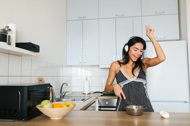 Aantrekkelijke jonge magere lachende vrouw plezier koken van eieren in de keuken in de ochtend ontbijten gekleed in pyjama's outfit, luisteren naar muziek op koptelefoon dansen