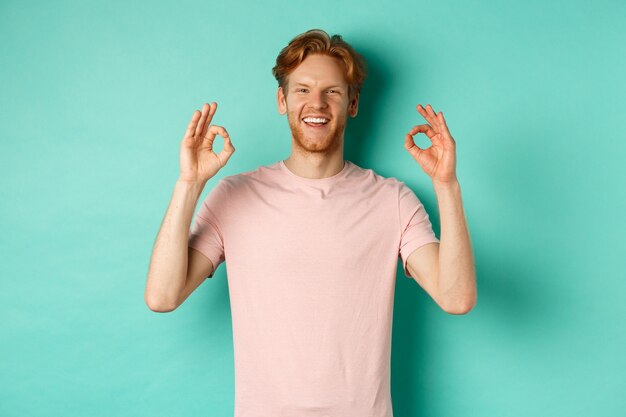 Aantrekkelijke jonge man in t-shirt die tevreden glimlacht, goedkeurend knikt en een OK-teken toont, goedkeurt en akkoord gaat met iets cools, staande over een turkooizen achtergrond.