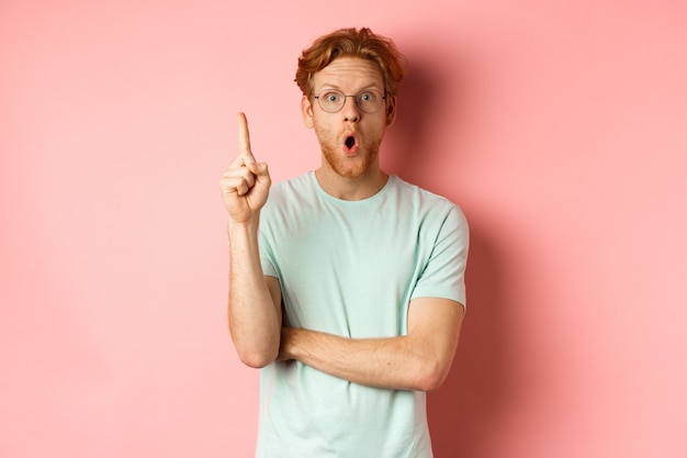 Gratis foto aantrekkelijke jonge man met rood haar, vinger opstekend in eureka-gebaar en werpend op idee, staande over roze achtergrond