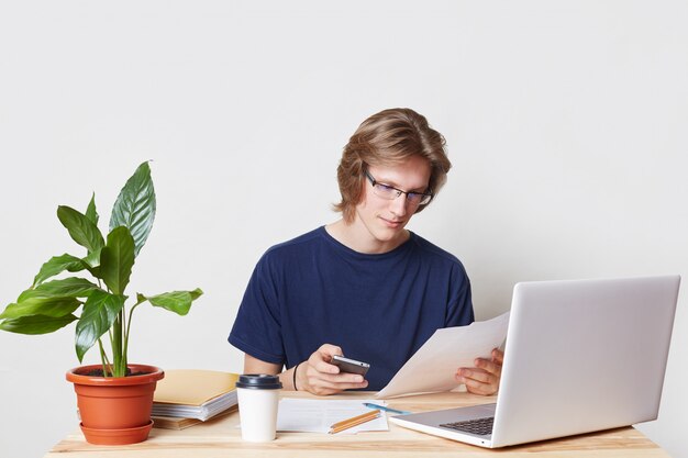 Aantrekkelijke jonge mannelijke ondernemer heeft serieuze uitstraling, sms-bericht op mobiele telefoon, maakt gebruik van gratis internetverbinding, doet papierwerk tijdens koffiepauze, geïsoleerd over witte blinde muur