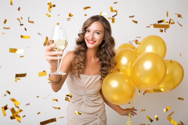 Aantrekkelijke jonge stijlvolle vrouw Nieuwjaar vieren, champagne drinken met luchtballons, gouden confetti vliegen, glimlachend gelukkig, wit, geïsoleerd, feestjurk dragen