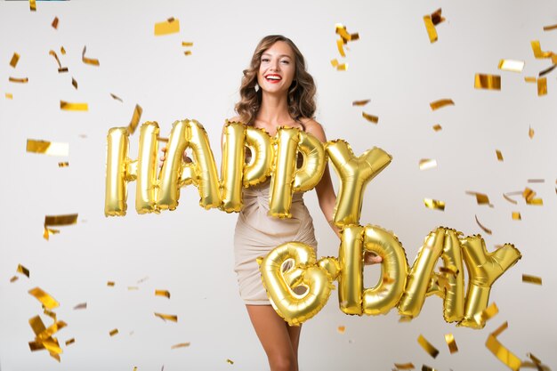 Aantrekkelijke jonge stijlvolle vrouw vieren, met lucht ballonnen gelukkige verjaardag brieven, gouden confetti vliegen, glimlachend gelukkig, geïsoleerd, feestjurk dragen