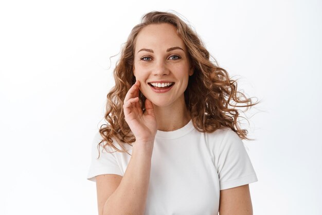 Aantrekkelijke jonge vrouw die het natuurlijke gezicht aanraakt zonder make-up, tevreden glimlacht en naar de camera kijkt die in een t-shirt staat tegen een witte achtergrond