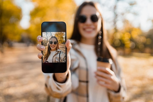 Aantrekkelijke jonge vrouw die in de herfst loopt en een jas draagt met behulp van de telefoon