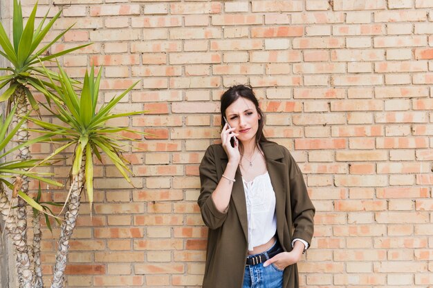 Aantrekkelijke jonge vrouw die op cellphone spreekt die zich tegen bakstenen muur bevindt