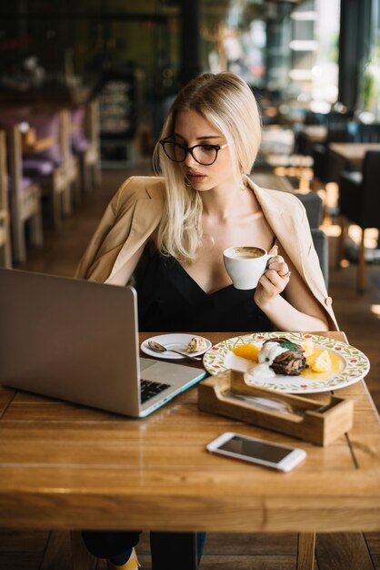 Aantrekkelijke jonge vrouw die op de koffiekop van de laptopholding werkt