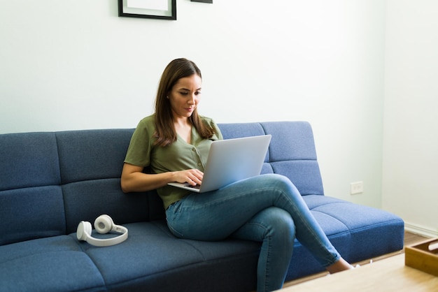 Aantrekkelijke jonge vrouw die op haar laptop typt terwijl ze op haar bank zit. Spaanse freelance werkt vanuit huis als bedrijfsmanager