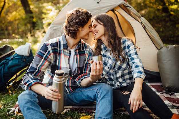 Aantrekkelijke jonge vrouw en knappe man brengen samen tijd door in de natuur. Zittend in een toeristische tent in het bos en koffie drinken.