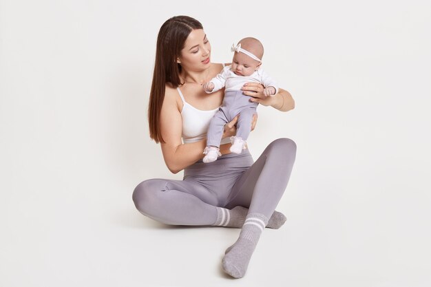 Aantrekkelijke jonge vrouw, gekleed in grijze legging en mouwloos t-shirt zittend op de vloer met pasgeboren kind, donkerharige vrouw draagt haar dochter, kind met haarband, geïsoleerd op een witte muur.