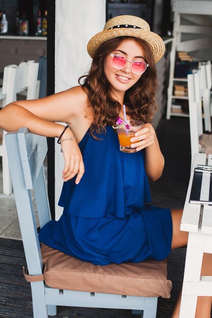 Aantrekkelijke jonge vrouw in blauwe jurk en stro hoed dragen roze zonnebril alcohol cocktails drinken op tropische vakantie zittend aan tafel in bar in zomer stijl outfit, glimlachend gelukkig in feeststemming