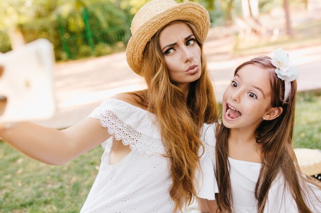 Aantrekkelijke jonge vrouw in kanten jurk gek rond met dochter voor foto, terwijl rust in park. Stijlvolle dame en schattig klein meisje maken grappige gezichten voor selfie.