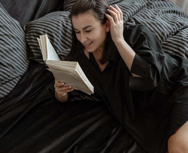 Aantrekkelijke jonge vrouw in pyjama's leest een boek terwijl ze in bed ligt.