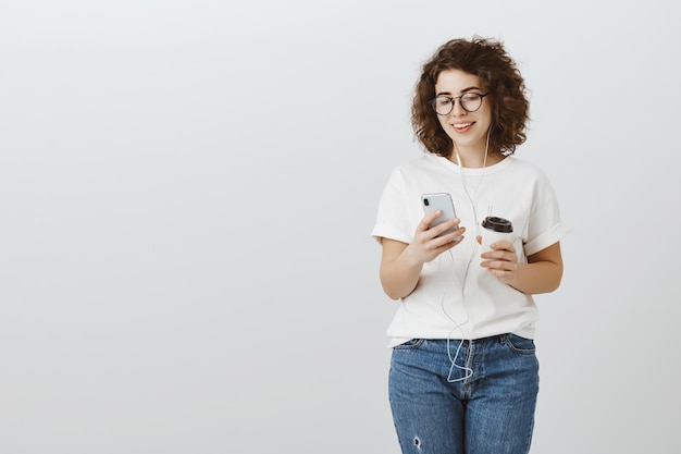 Aantrekkelijke jonge vrouw koffie drinken, messaging via mobiele telefoon en muziek in oortelefoons luisteren