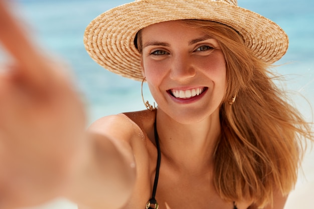 Aantrekkelijke jonge vrouw met brede glimlach, gezonde huid, rust aan kust, neemt foto van zichzelf, in goed humeur, geniet van vrije tijd en zomervakantie. Mooie vrouw maakt selfie tegen Oceaan