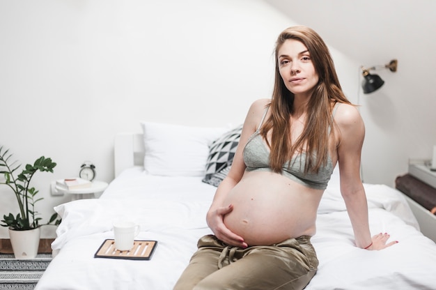 Gratis foto aantrekkelijke jonge zwangere vrouw zittend op bed haar buik aan te raken