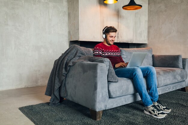 Aantrekkelijke jongeman op sofa thuis in de winter met smartphone in koptelefoon, luisteren naar muziek, dragen rode gebreide trui, bezig met laptop, freelancer