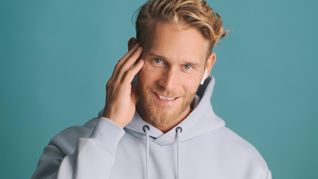 Aantrekkelijke lachende blonde bebaarde man in hoodie op zoek gelukkig luisteren muziek in draadloze koptelefoon geïsoleerd op blauwe achtergrond