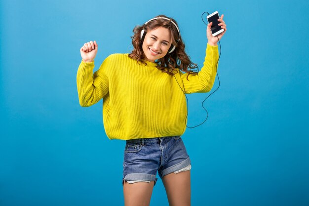 Aantrekkelijke lachende gelukkige vrouw dansen luisteren naar muziek in hoofdtelefoons in stijlvolle hipster outfit geïsoleerd op blauwe studio achtergrond, korte broek en gele trui dragen
