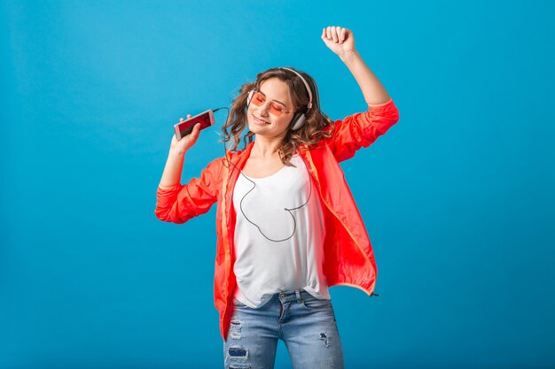 Aantrekkelijke lachende gelukkige vrouw dansen luisteren naar muziek in koptelefoon gekleed in hipster stijl outfit geïsoleerd op blauwe studio achtergrond, roze jas en zonnebril dragen