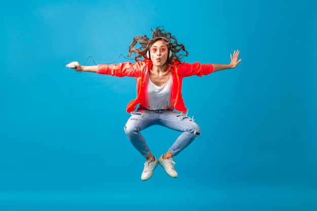 Aantrekkelijke lachende gelukkige vrouw dansen luisteren naar muziek in koptelefoon gekleed in hipster stijl outfit geïsoleerd op blauwe studio achtergrond, roze jas en zonnebril dragen