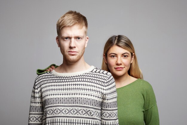 Aantrekkelijke lachende jonge vrouw met blond haar die achter ernstige ongeschoren man staat, hand op zijn schouder houdt als teken van steun, staande tegen grijze muur met kopie ruimte voor uw informatie