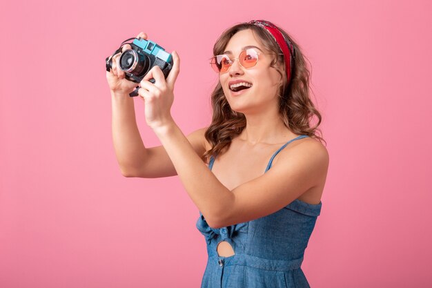 Aantrekkelijke lachende vrouw die foto op vintage camera draagt denim jurk en zonnebril, geïsoleerd op roze achtergrond