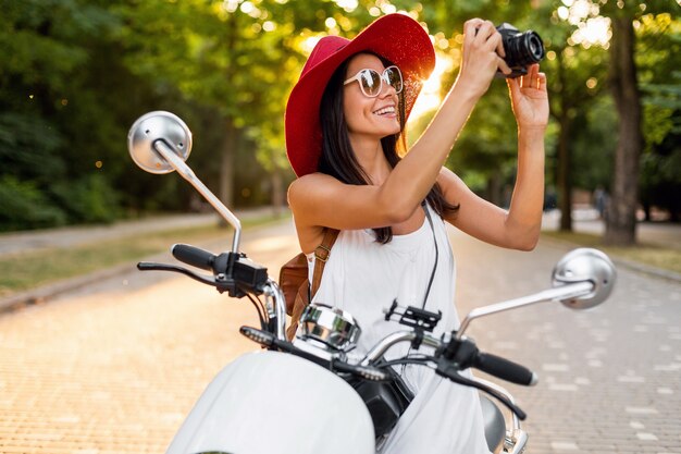 Aantrekkelijke lachende vrouw rijden op motor in straat in zomer stijl outfit dragen witte jurk en rode hoed reizen op vakantie, fotograferen op vintage fotocamera