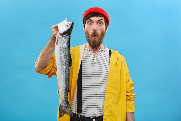 Gratis foto aantrekkelijke man met baard gekleed in rode hoed, gele regenjas en overall met enorme vis kijken met afgeluisterde ogen en geopende mond met een schok die zulke grote vissen niet eerder vangen