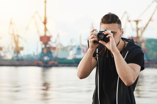 Aantrekkelijke man werken met camera. Jonge stijlvolle fotograaf kijkt door de camera tijdens fotosessie met prachtig model, het nemen van foto's in de haven in de buurt van kust, met de nadruk op werk