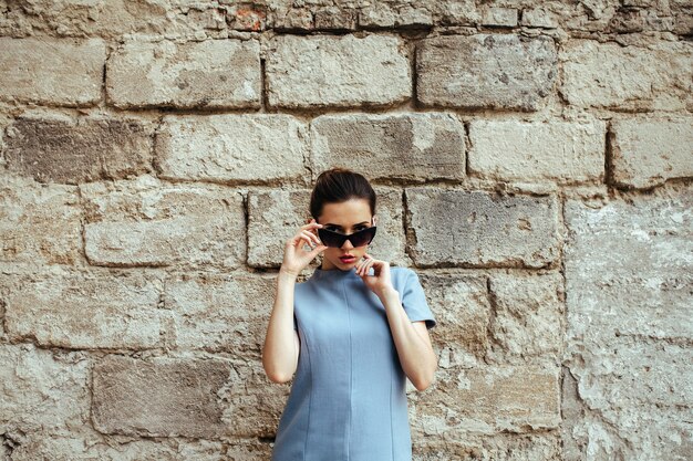 Aantrekkelijke mode vrouw in blauwe jurk met zonnebril poseren in de buurt van witte muur