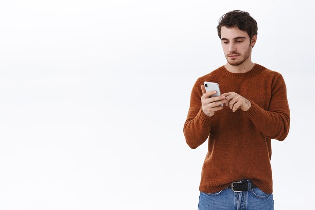 Aantrekkelijke moderne jonge man met baard in sweatshirt casual outfit met behulp van mobiele telefoon tik smartphone scherm bericht sms'en of browsen internet scroll dating app feed witte achtergrond