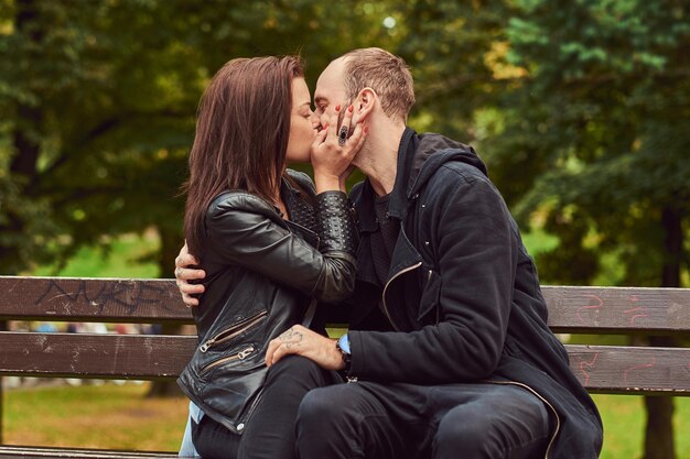 Aantrekkelijke moderne paar zoenen op een date terwijl ze knuffelen op een bankje in het park. Genieten van hun liefde en de natuur.