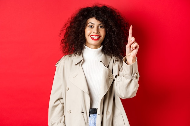 Aantrekkelijke moderne vrouw met krullend kapsel met rode lippen, lente-trenchcoat, wijzende vinger omhoog...