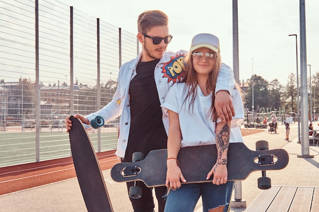 Aantrekkelijke paar trendy geklede jonge hipsters poseren met skateboards in het stadssportcomplex op zonnige dag, met warme tonen.