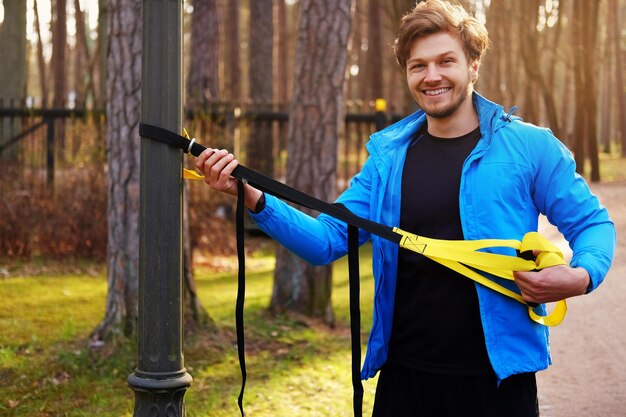 Aantrekkelijke positieve man in een blauwe regenjas die traint in een park met trx-fitnessstrips.