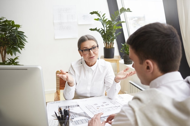 Aantrekkelijke rijpe vrouwelijke CEO in glazen en wit overhemd zittend op haar werkplek en jonge getalenteerde mannelijke sollicitant voor chief atchitect-positie interviewen, hem vragen over werkervaring en vaardigheden