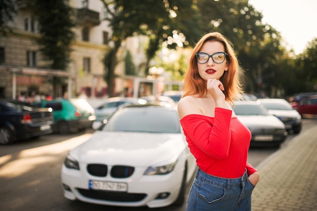 Gratis foto aantrekkelijke roodharige vrouw met een bril draagt een rode blouse en jeansrok die zich op straat tegen een witte sportwagen