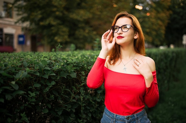 Aantrekkelijke roodharige vrouw met een bril draagt een rode blouse en jeansrok die zich voordeed in het groene park
