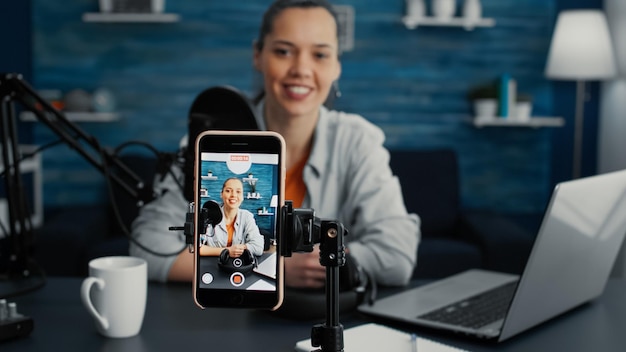 Aantrekkelijke social media influencer die aan het bureau van de thuisstudio zit terwijl hij begint te streamen vanuit de woonkamer met smartphone. Jonge contentmaker die dagelijkse vlog opneemt met modern touchscreen-apparaat.