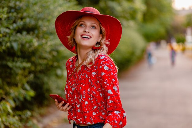 Aantrekkelijke stijlvolle blonde glimlachende vrouw in rode strooien hoed en blouse zomer mode outfit met behulp van telefoon