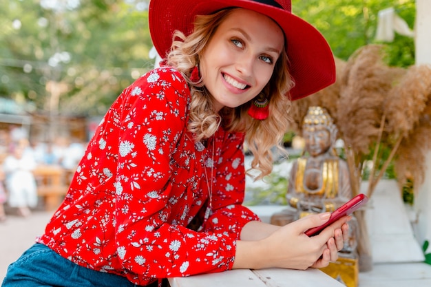 Aantrekkelijke stijlvolle blonde lachende vrouw in stro rode hoed en blouse zomer mode outfit bedrijf met behulp van slimme telefoon café