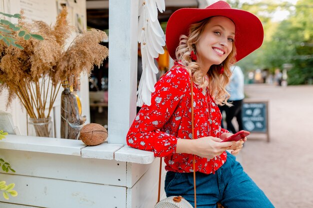 Aantrekkelijke stijlvolle blonde lachende vrouw in stro rode hoed en blouse zomer mode outfit bedrijf met behulp van slimme telefoon café