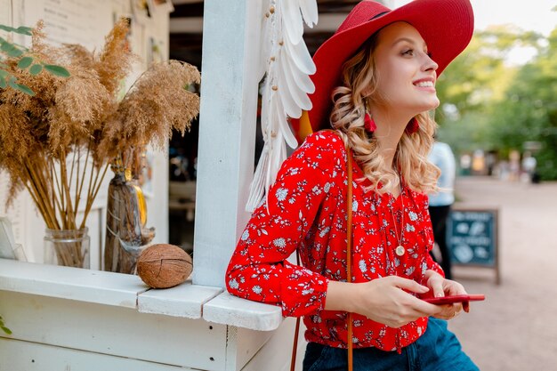 Aantrekkelijke stijlvolle blonde lachende vrouw in stro rode hoed en blouse zomer mode outfit bedrijf met behulp van slimme telefoon café