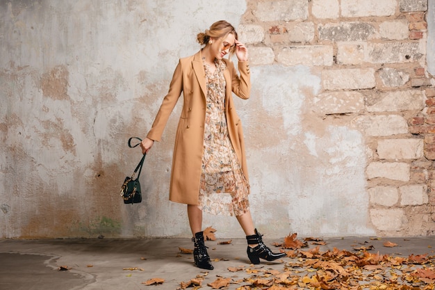 Gratis foto aantrekkelijke stijlvolle blonde vrouw in beige jas wandelen in straat tegen vintage muur