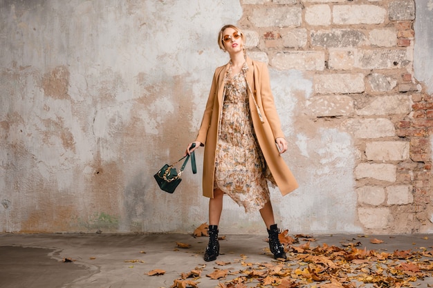 Aantrekkelijke stijlvolle blonde vrouw in beige jas wandelen in straat tegen vintage muur