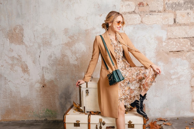 Aantrekkelijke stijlvolle blonde vrouw in beige jas zittend op koffers tegen muur in straat
